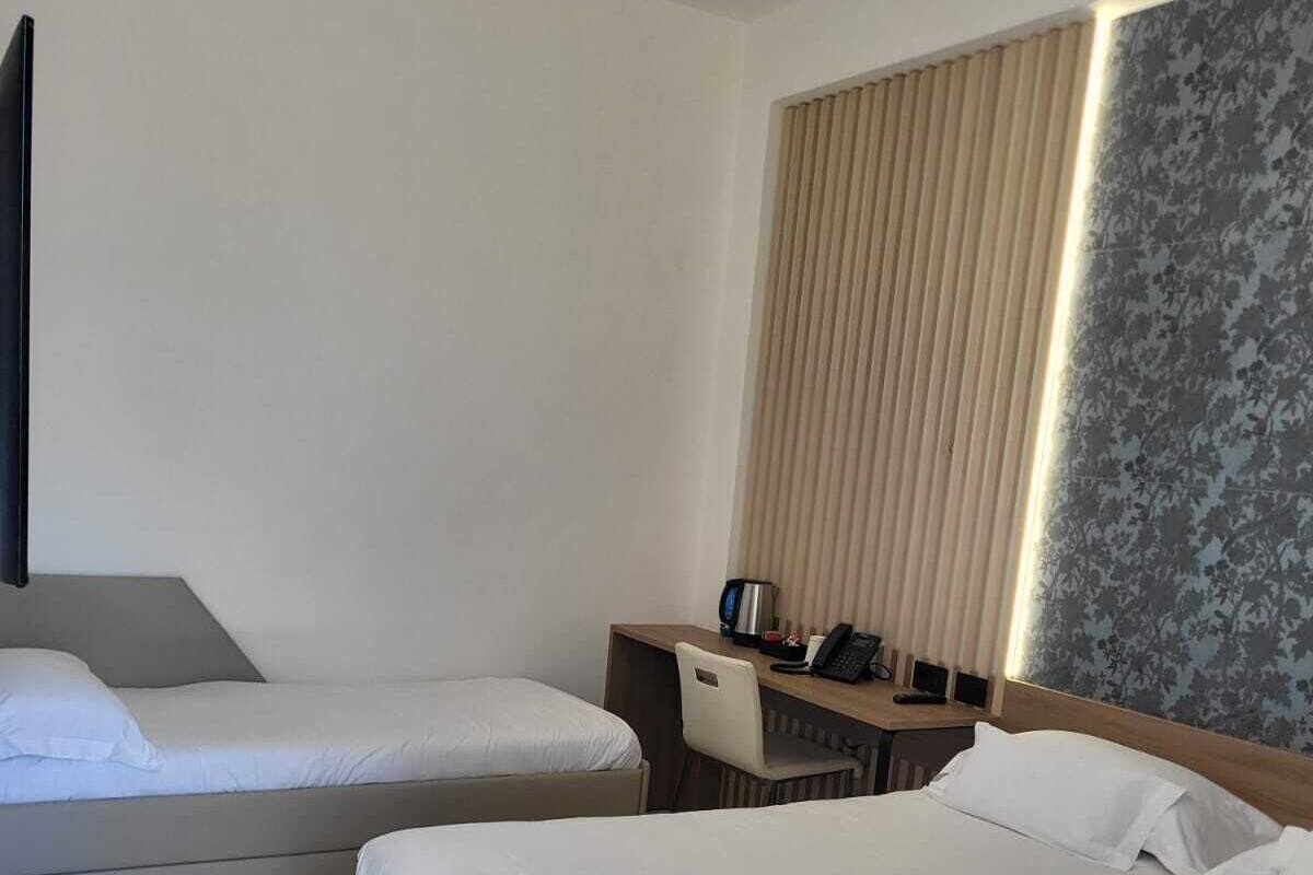 Une chambre d’hôtel avec un grand lit, un lit plus petit, un bureau avec une chaise, un téléphone et une fenêtre avec des stores.