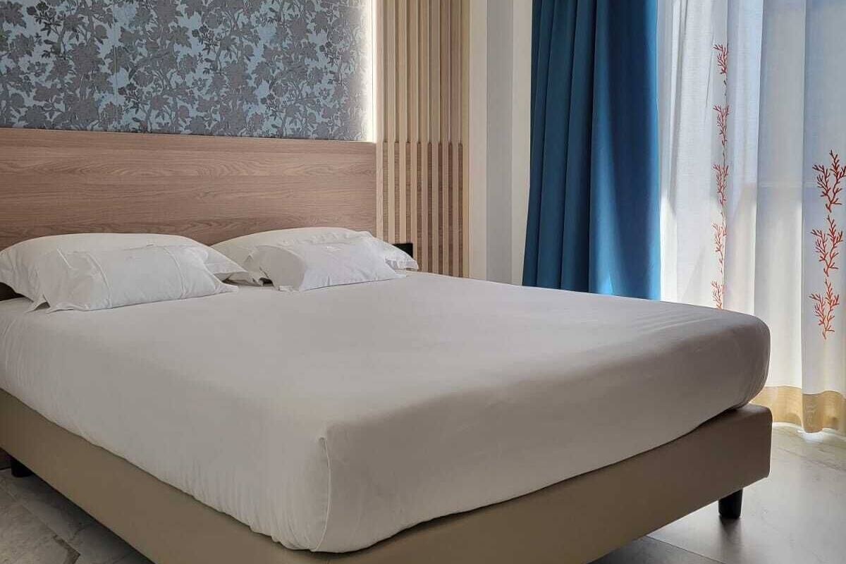 Un grand lit avec une tête de lit en bois, un sol carrelé de marbre et une vue sur l’extérieur à travers de fines rideaux.