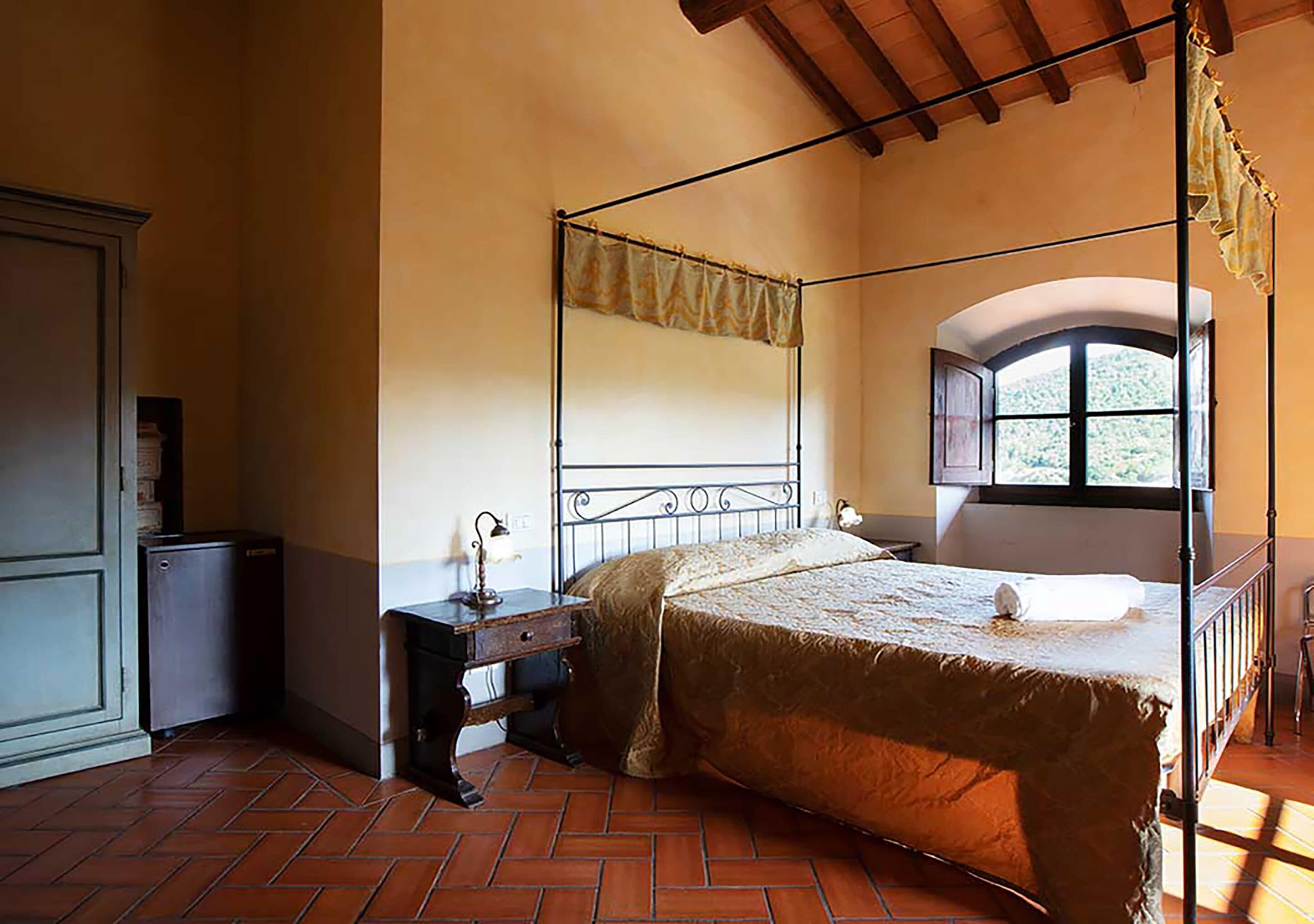 A bedroom with a four-poster bed, a small bedside table, a wardrobe, and a window with a view of greenery.