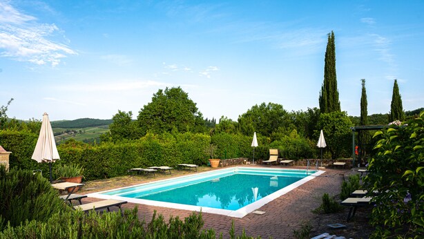 A rectangular swimming pool with a clear blue water surface, surrounded by a brick patio. There are several lounge chairs and umbrellas around the pool area. - Villa Rosa Panzano (Greve in Chianti)