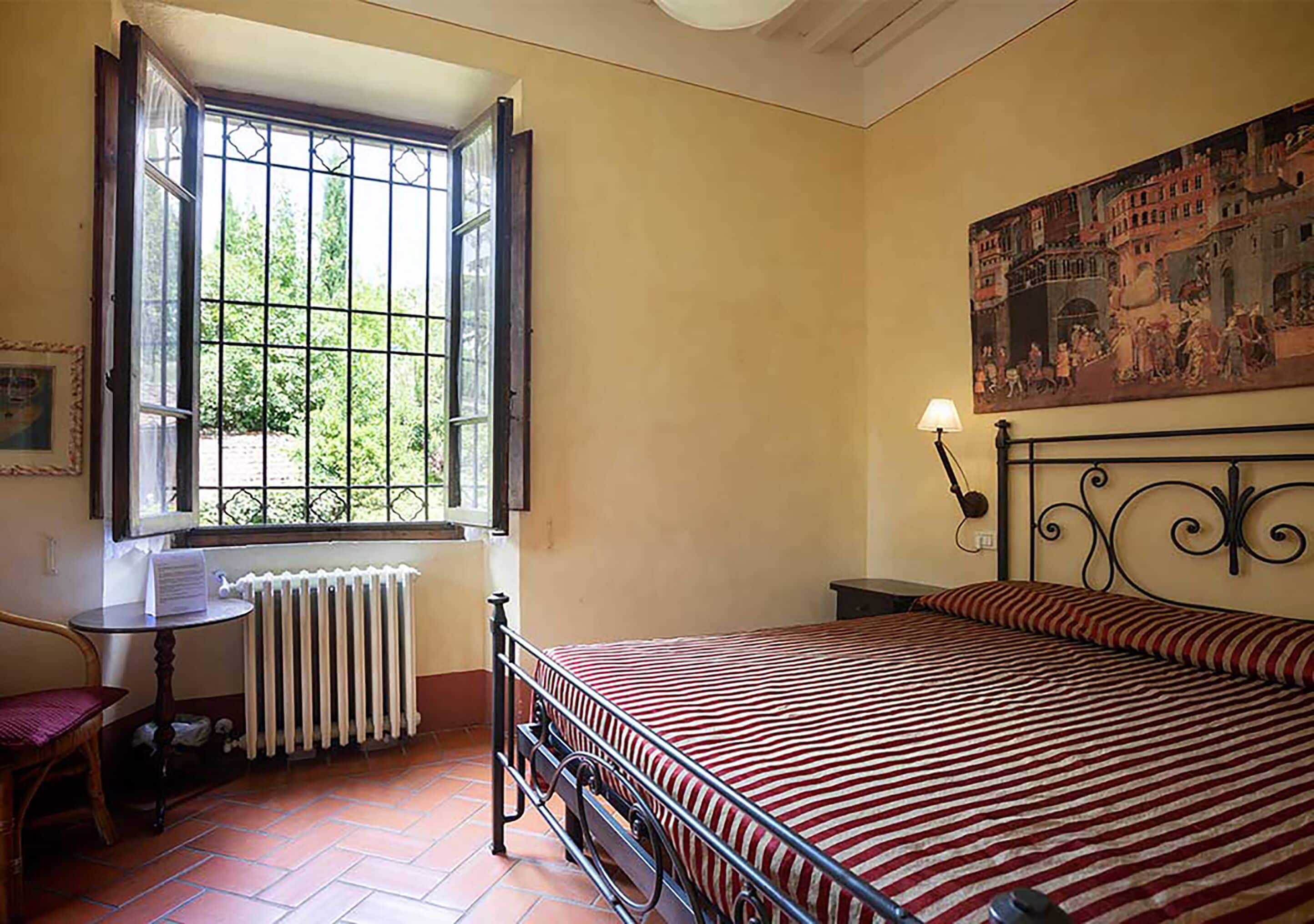 Une chambre à coucher avec un lit, un radiateur, une fenêtre dont les volets sont ouverts, une chaise et un tableau accroché au mur.