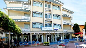 A hotel with a sign reading "Hotel Maxim," featuring a multi-story structure with balconies and a covered outdoor seating area with blue chairs and tables. - Hotel Maxim (Caorle)