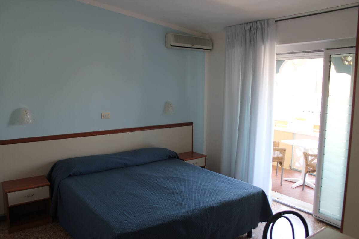 A bedroom with a bed, a nightstand, a drawer, an air conditioning unit, and a view of an outdoor area with chairs and a table.