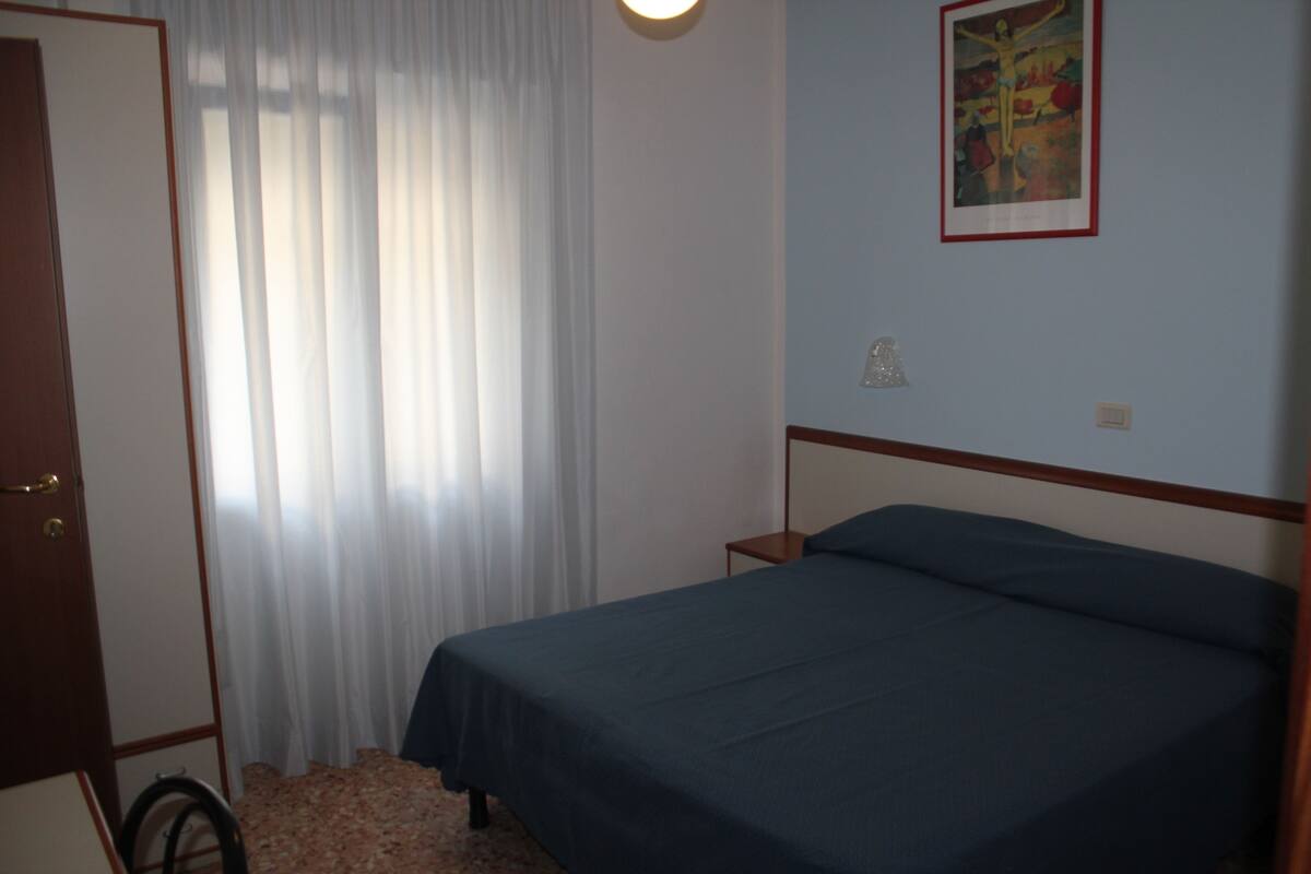A bedroom with a bed, a nightstand, a lamp, a window with curtains, and a painting on the wall.