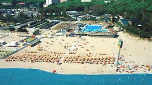 An aerial view of a beach with numerous sunbeds and umbrellas, a swimming area, and a building with a blue roof. - Sole (Rosolina)