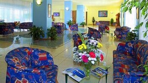A hotel lobby with colorful patterned sofas, a central floral arrangement, and a large window with natural light. - Sole (Rosolina)