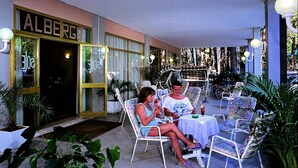 An outdoor seating area with tables and chairs, a building entrance with a sign reading "ALBERC", and potted plants. - Sole (Rosolina)