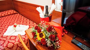 Una habitación de hotel con una cama, una mesita de noche de madera, un ramo de rosas rojas, una botella de champán y un control remoto.