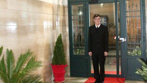 Un hombre de pie sobre una alfombra roja frente a un edificio con puerta de vidrio y una planta en maceta a la izquierda.