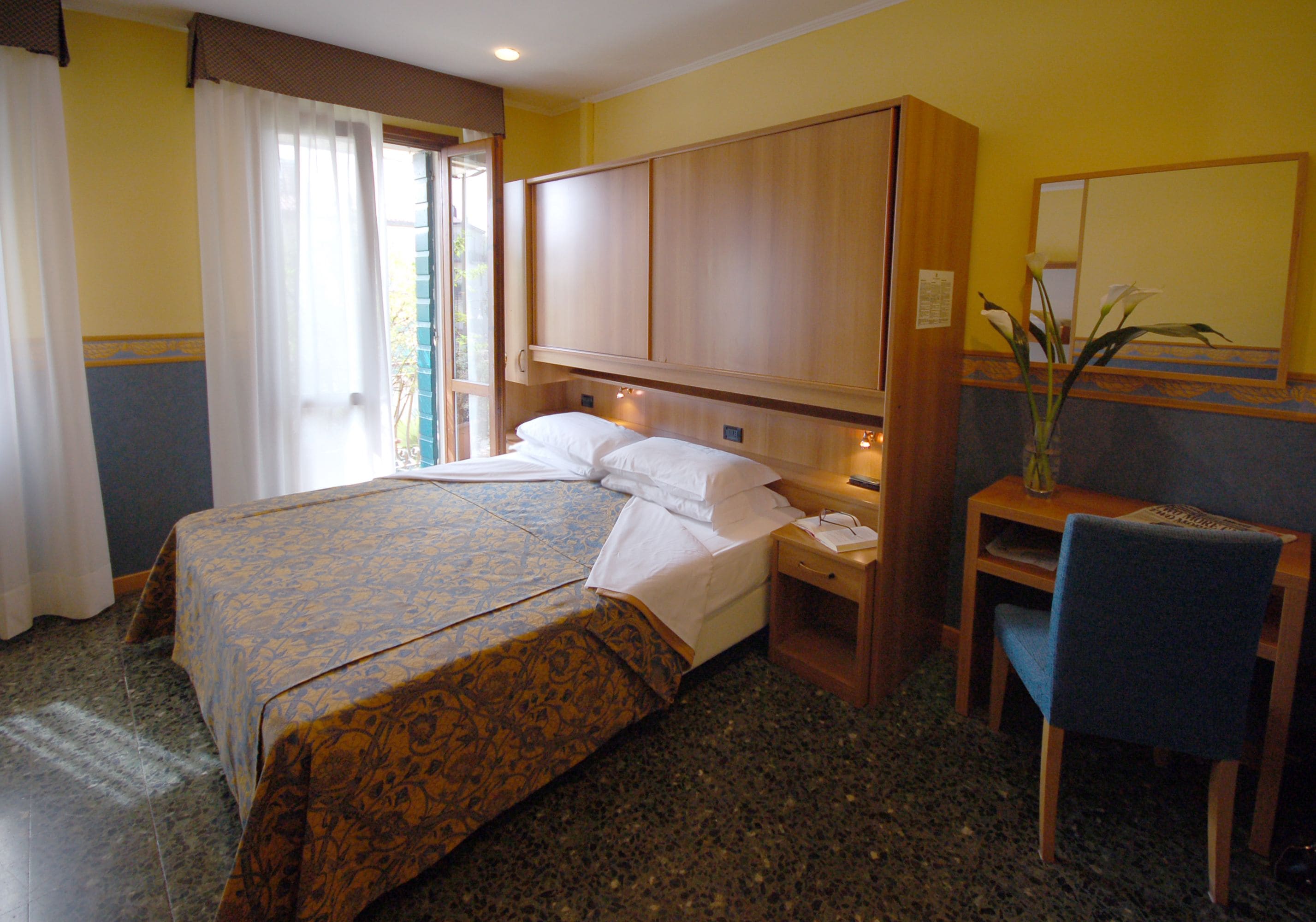 A hotel room with a large bed, a wooden headboard, a desk with a chair, and a mirror on the wall.