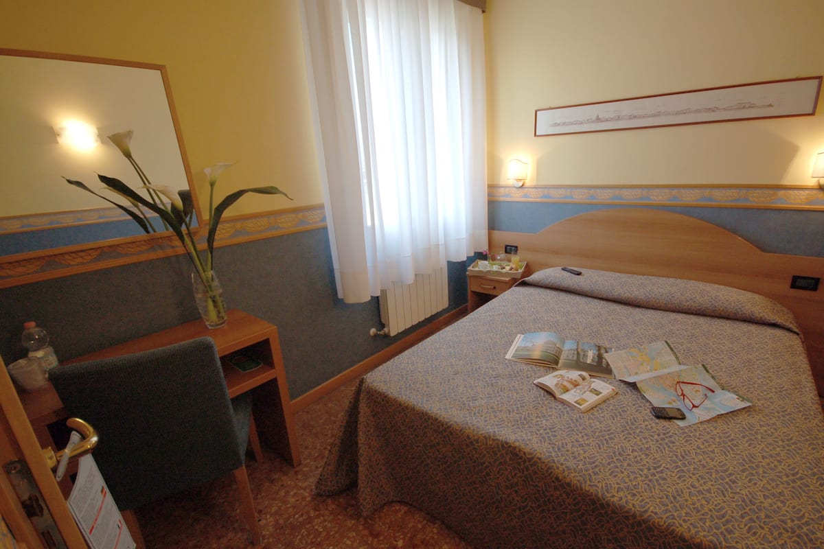 Habitación de hotel con cama, escritorio con silla, florero con flores, ventana con cortinas y una lámpara de pared.