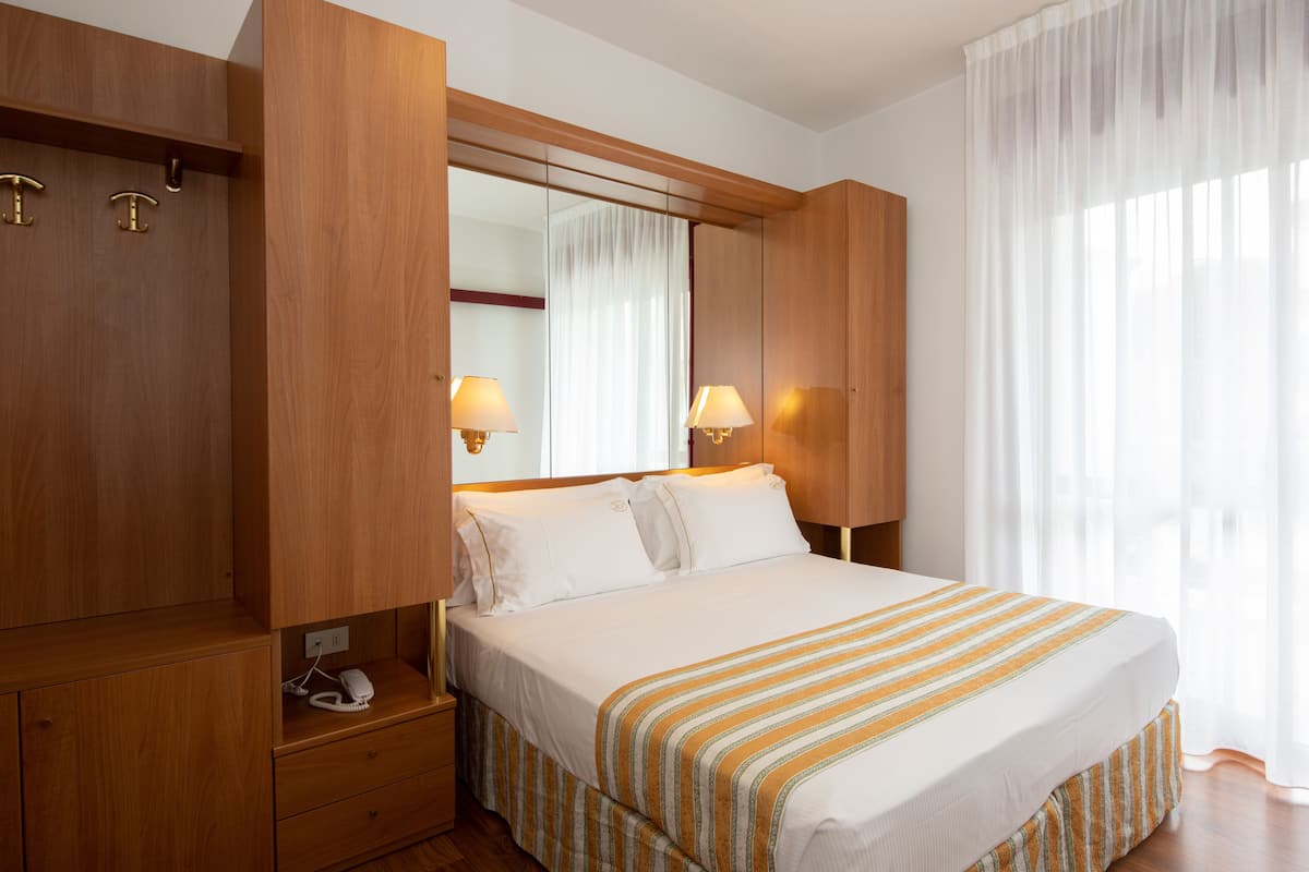 A hotel room with a large bed, wooden wardrobe, bedside lamps, and a large window with sheer curtains.