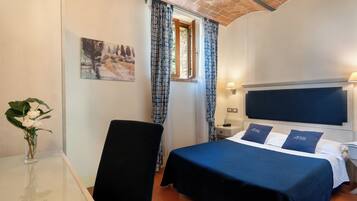 Habitación de hotel con cama, una silla, una mesita con un florero y cortinas en la ventana, además de un cuadro en la pared.