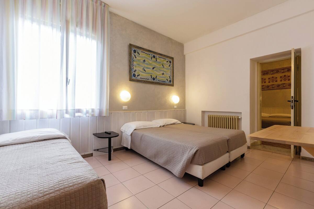 A hotel room with two beds, a wooden table, a radiator, and a painting on the wall.