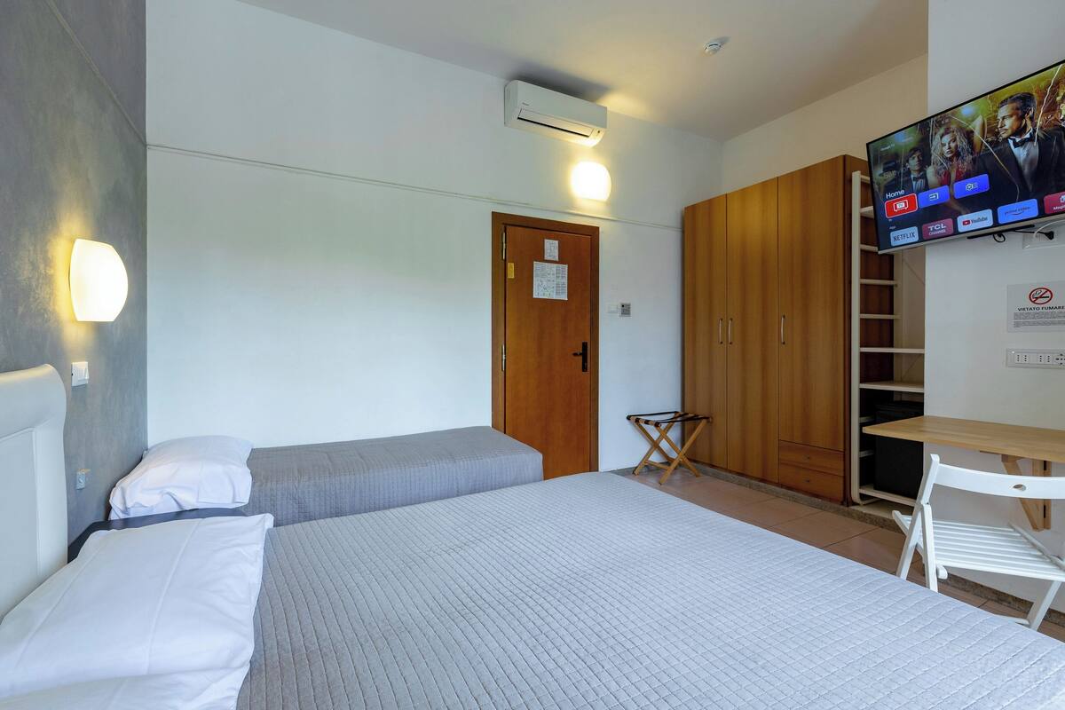A hotel room with two beds, a TV mounted on the wall, a wooden wardrobe, and a small table with a chair.