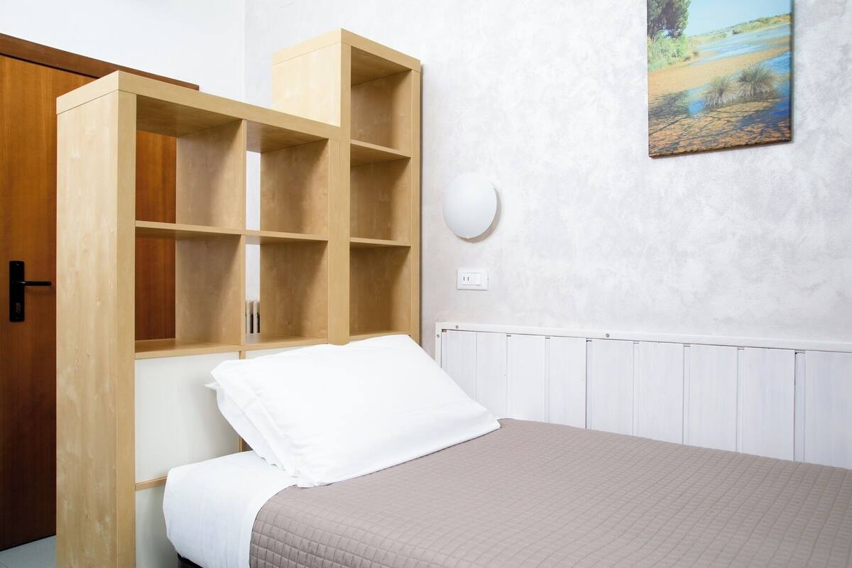 A bedroom with a bed, a wooden wardrobe, a white wall, a painting, and a white radiator.