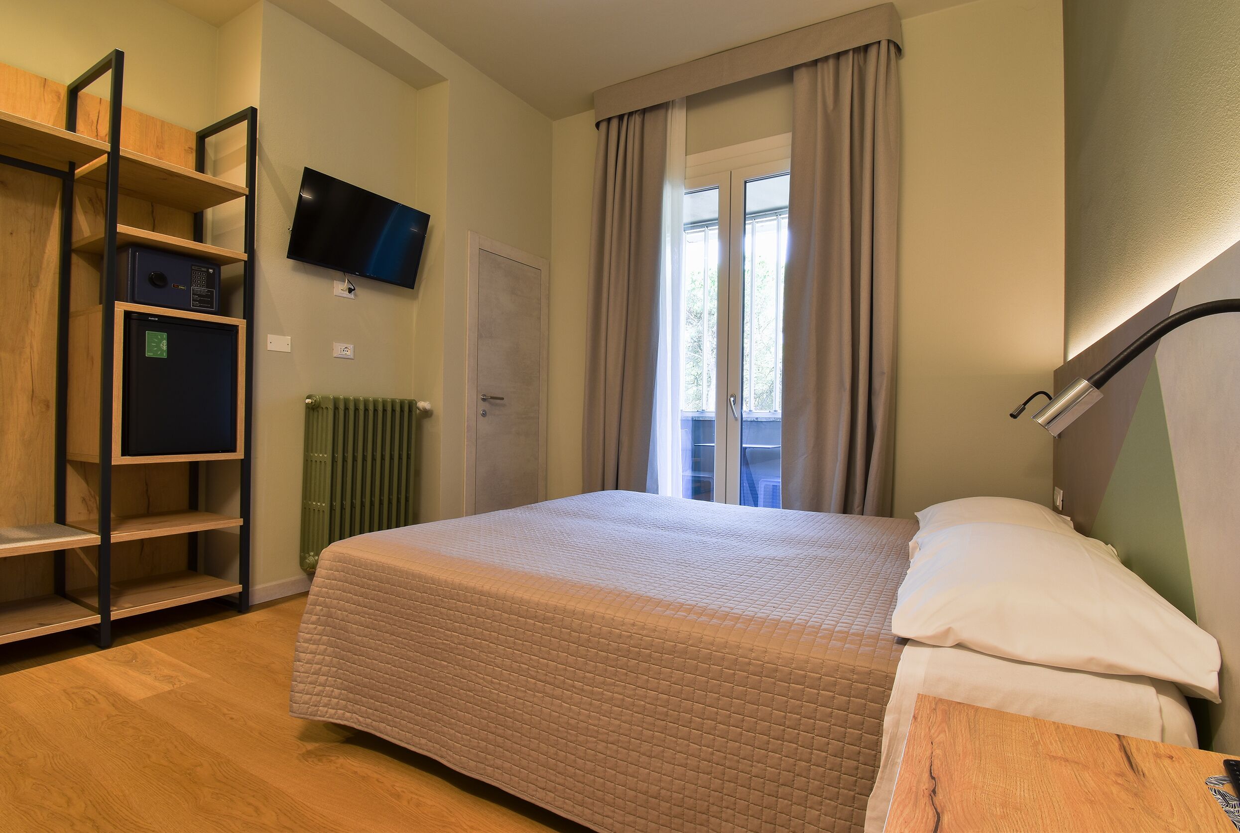 Un dormitorio con una cama, un televisor montado en la pared, una estantería de madera y una ventana con cortinas.