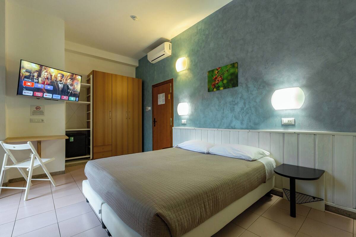 A bedroom with a bed, a desk, a chair, a TV, and a wall-mounted air conditioner.