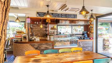 Interior de un restaurante con una mesa de madera y sillas coloridas, una barra con una máquina de café y un menú en una pizarra.