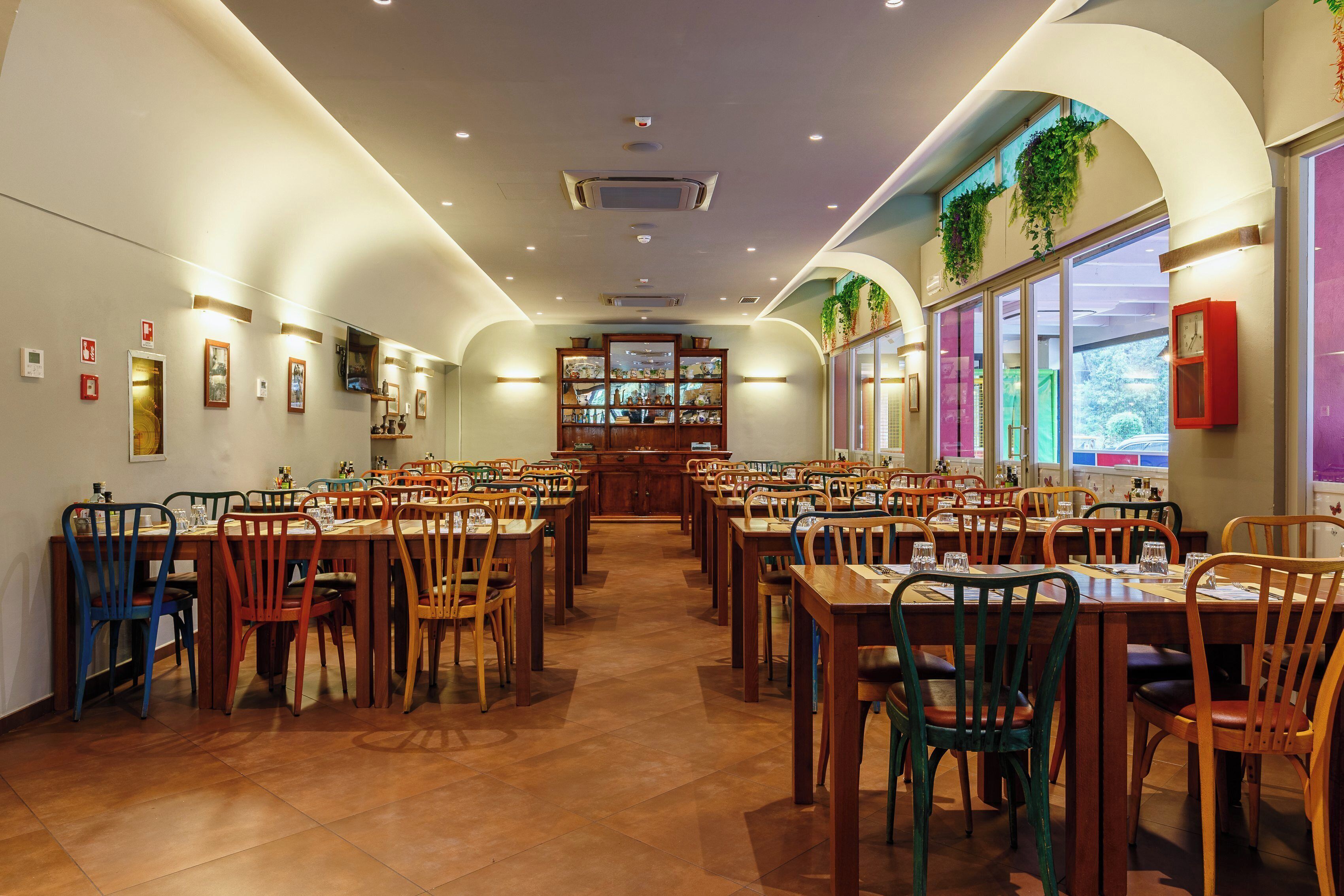 Un'ampia sala da ristorante con diversi tavoli e sedie, una vetrina centrale e grandi finestre con piante.