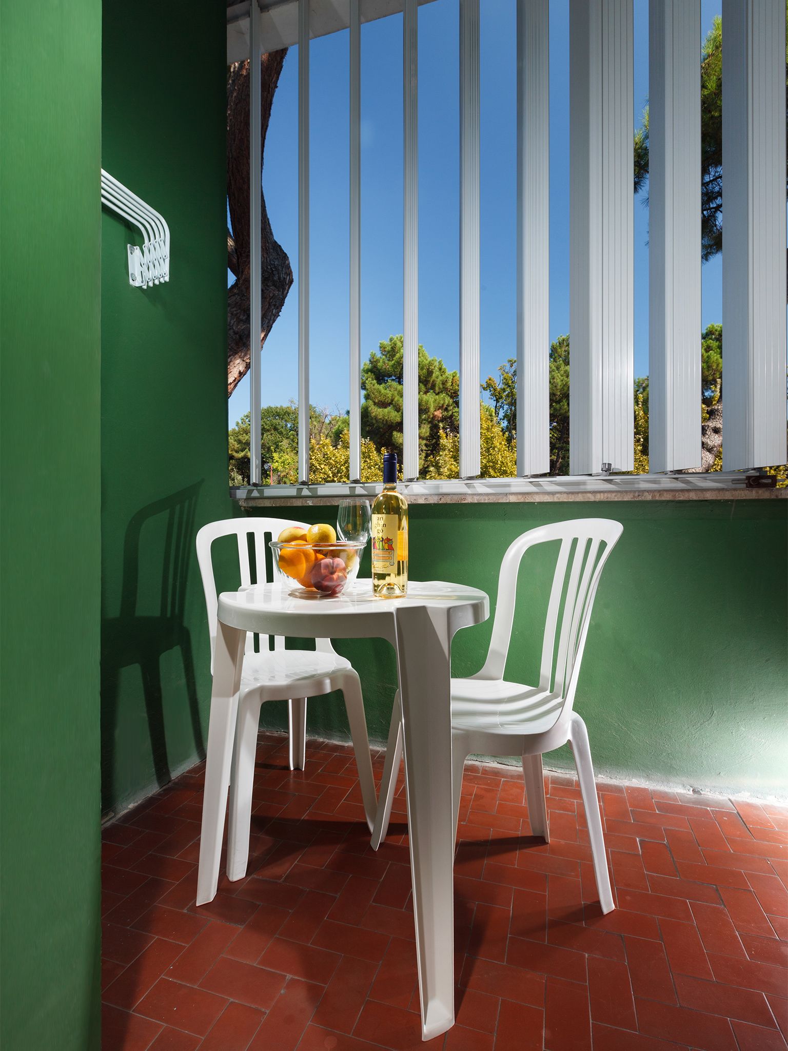 A small outdoor seating area with a table and two chairs, a bowl of fruit, and a bottle of wine.