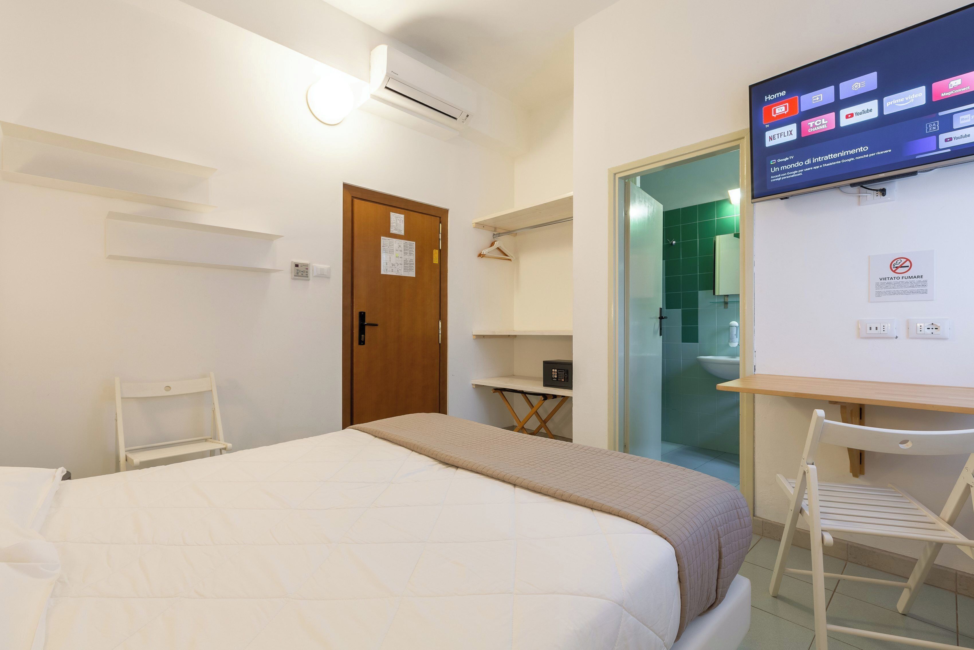 A hotel room with a bed, a desk, a chair, a TV mounted on the wall, and a door.