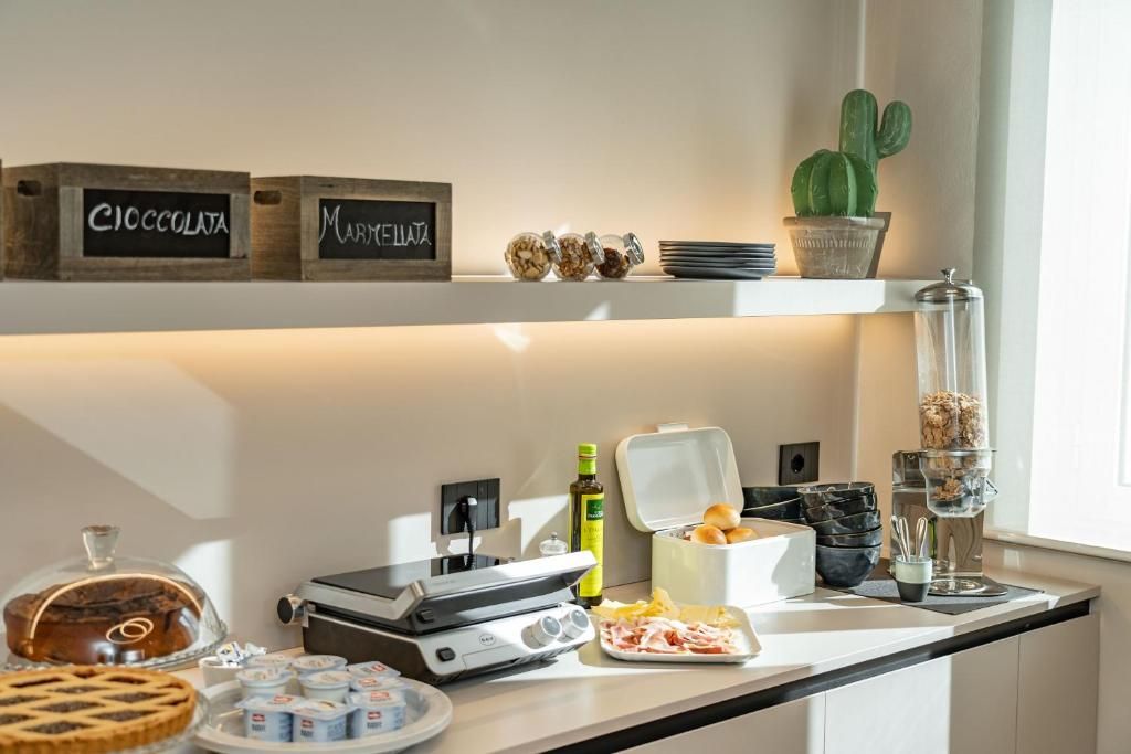 A kitchen counter with a waffle maker, a pie, and various food items. There are jars labeled 'CIOCCOLATA' and 'MARMELLATA' on the shelf above.