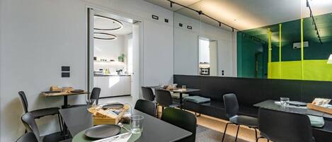 Un comedor moderno con mesas y sillas, una cocina al fondo y un espejo grande que refleja el espacio.