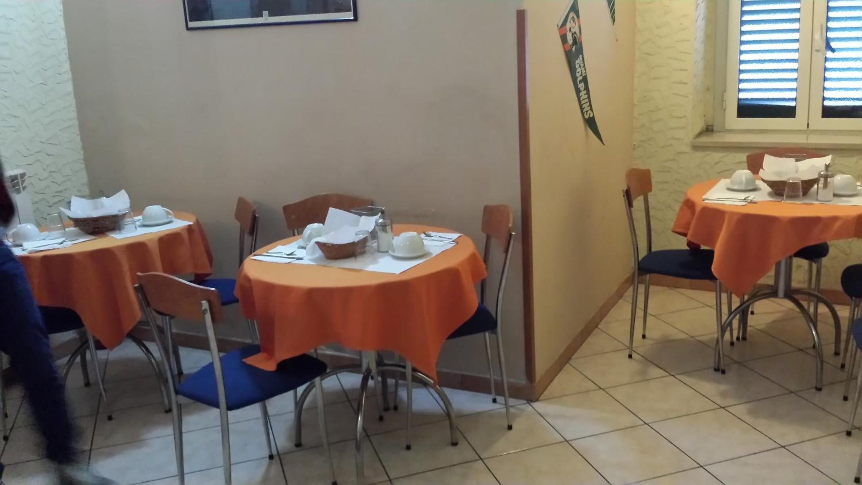 Zona pranzo con tavoli ricoperti da tovaglie arancioni, sedie e un cartello appeso al muro.