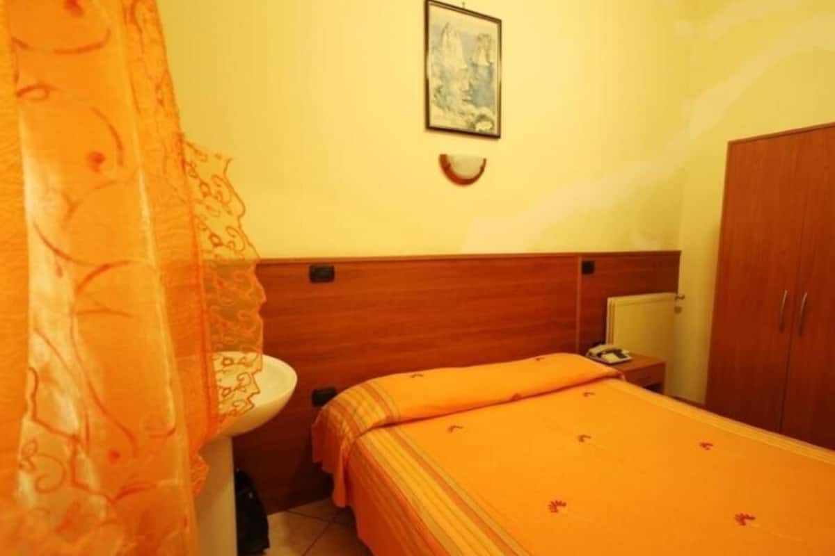 A bedroom with a bed, a wooden headboard, a bedside table, a nightstand, a wall-mounted light, and a painting.