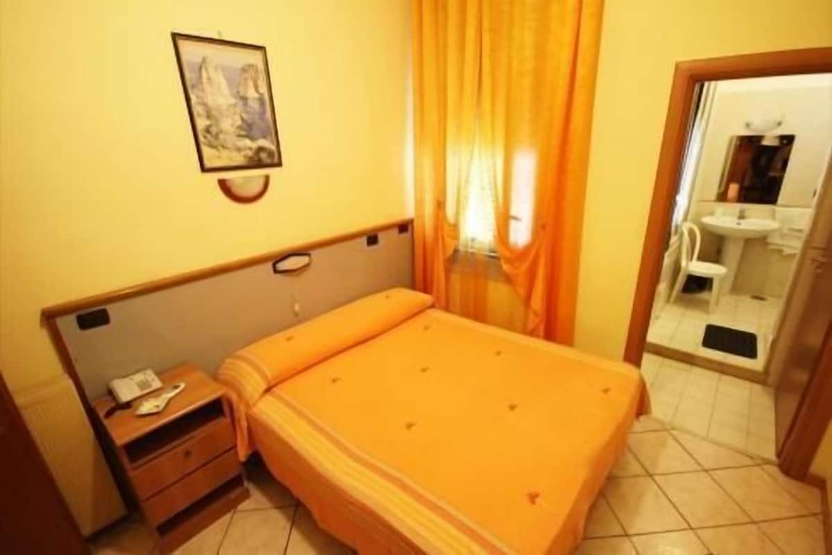 A bedroom with a bed, a nightstand, a painting on the wall, and a bathroom visible through an open door.