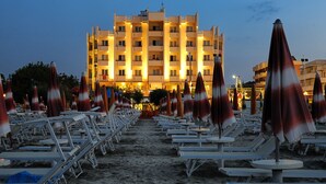 A beachside hotel with rows of sun loungers and umbrellas, a large building in the background, and a sandy walkway leading to the beach. - Hotel Life (Rimini)