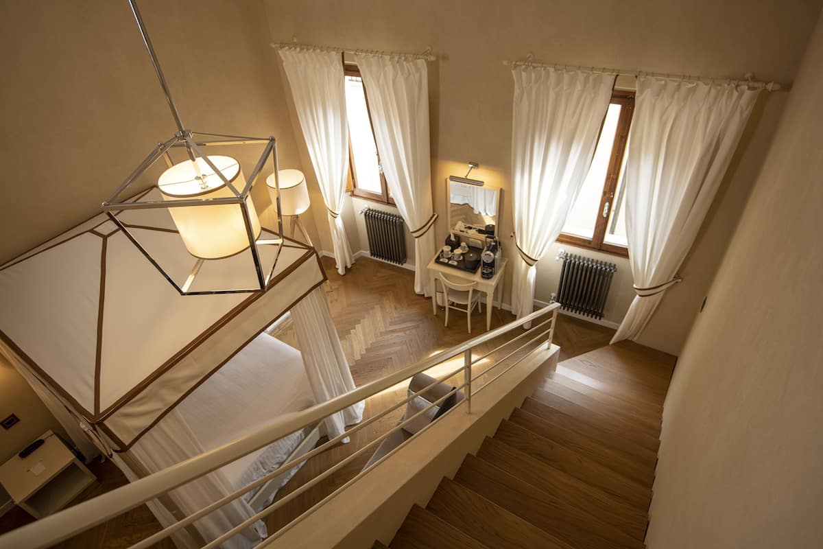 A bedroom with a wooden floor, a bed with white bedding, a nightstand, a dresser, a window with white curtains, and a staircase with a railing.