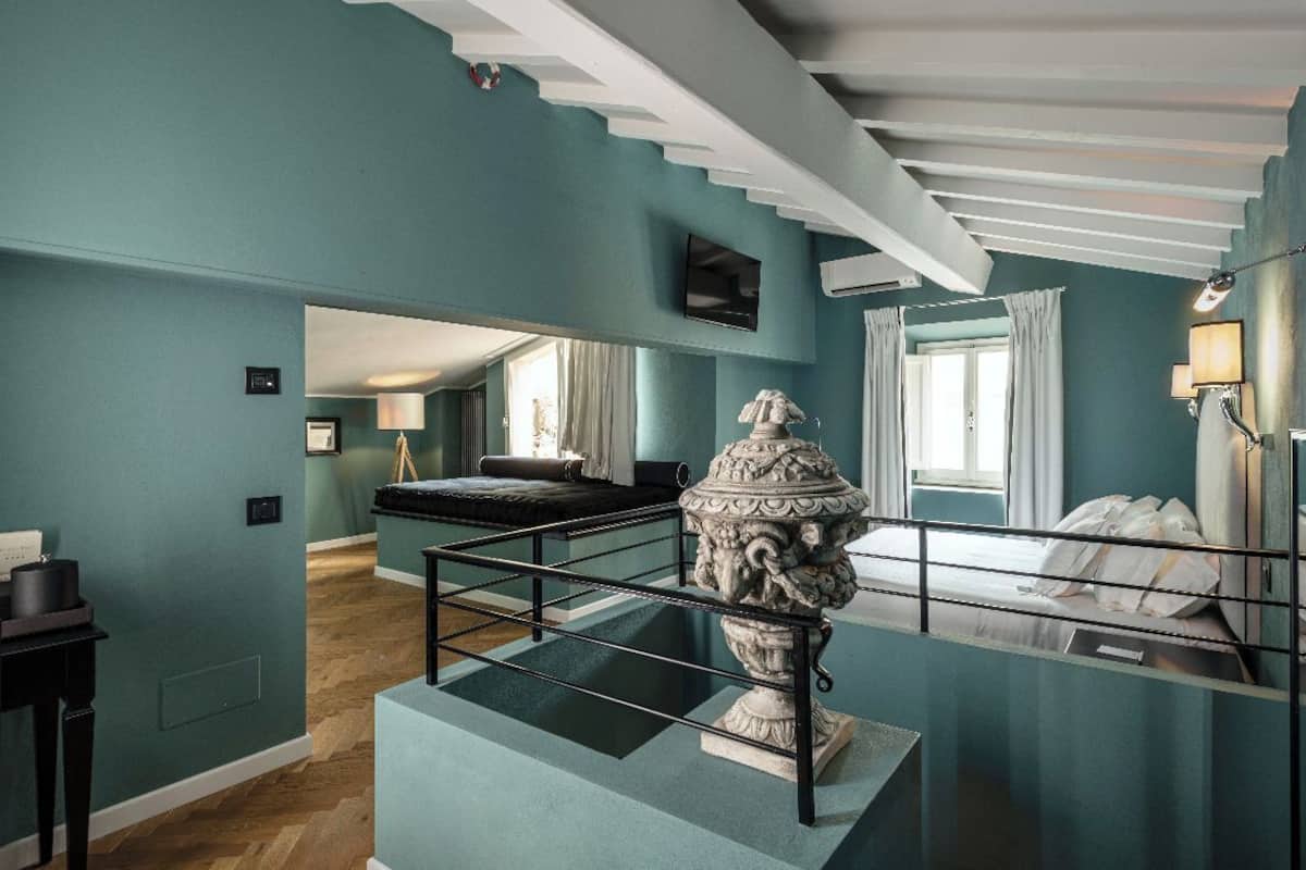 A room with teal walls, a bed with white linens, a stone urn, a black table, and a mirror reflecting the room.