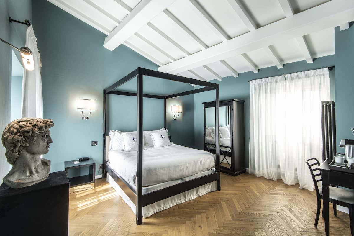 A four-poster bed in a room with a herringbone floor pattern, a bust sculpture on a pedestal, and a desk with a chair.