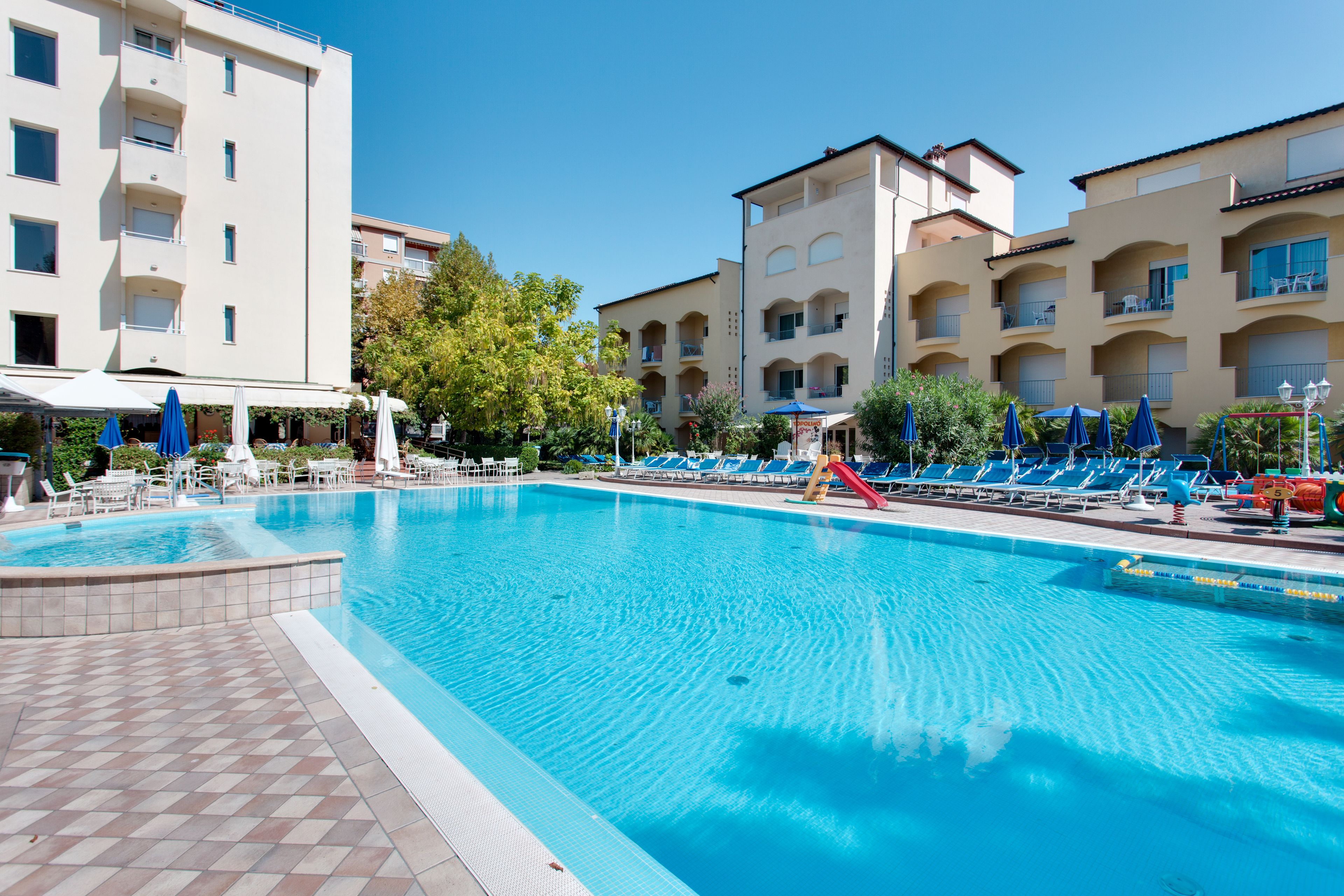 Una piscina con lettini e ombrelloni, circondata da edifici a più piani con balconi.