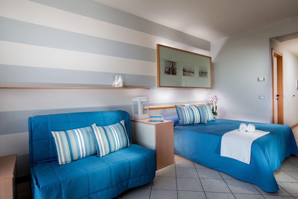 A hotel room with two blue beds, a wooden nightstand, and a framed picture on the wall.