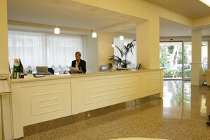 Ein Empfangsbereich mit Rezeptionist, einem Schreibtisch mit Computer und einem großen Fenster mit Vorhängen.
