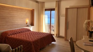 A hotel room with a bed, a desk, a chair, a window with curtains, and a view of the sea. - Palazzina (Gargnano)