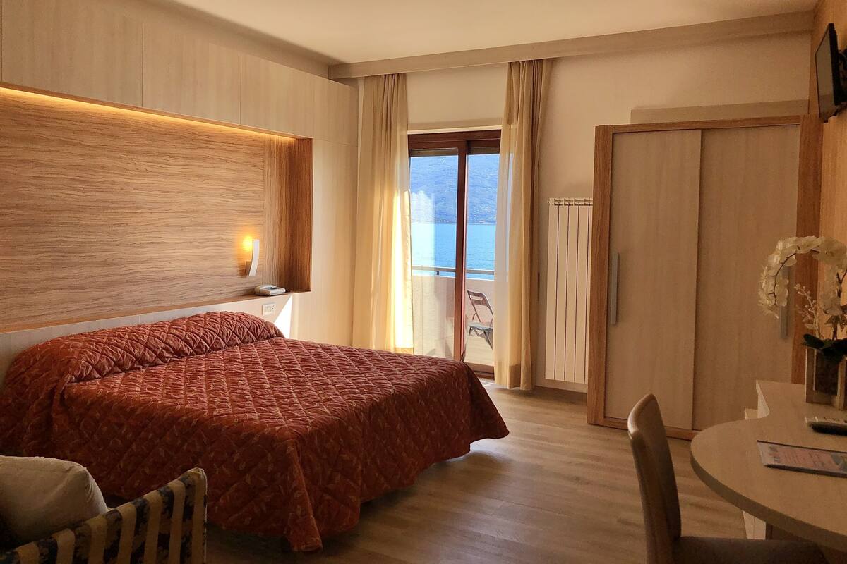 A hotel room with a bed, a desk, a chair, a window with curtains, and a view of the sea.