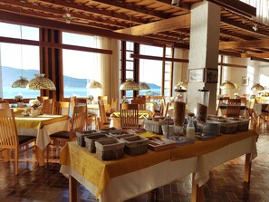 A dining area with tables set for breakfast, wooden beams on the ceiling, and large windows with a view of the sea. - Palazzina (Gargnano)