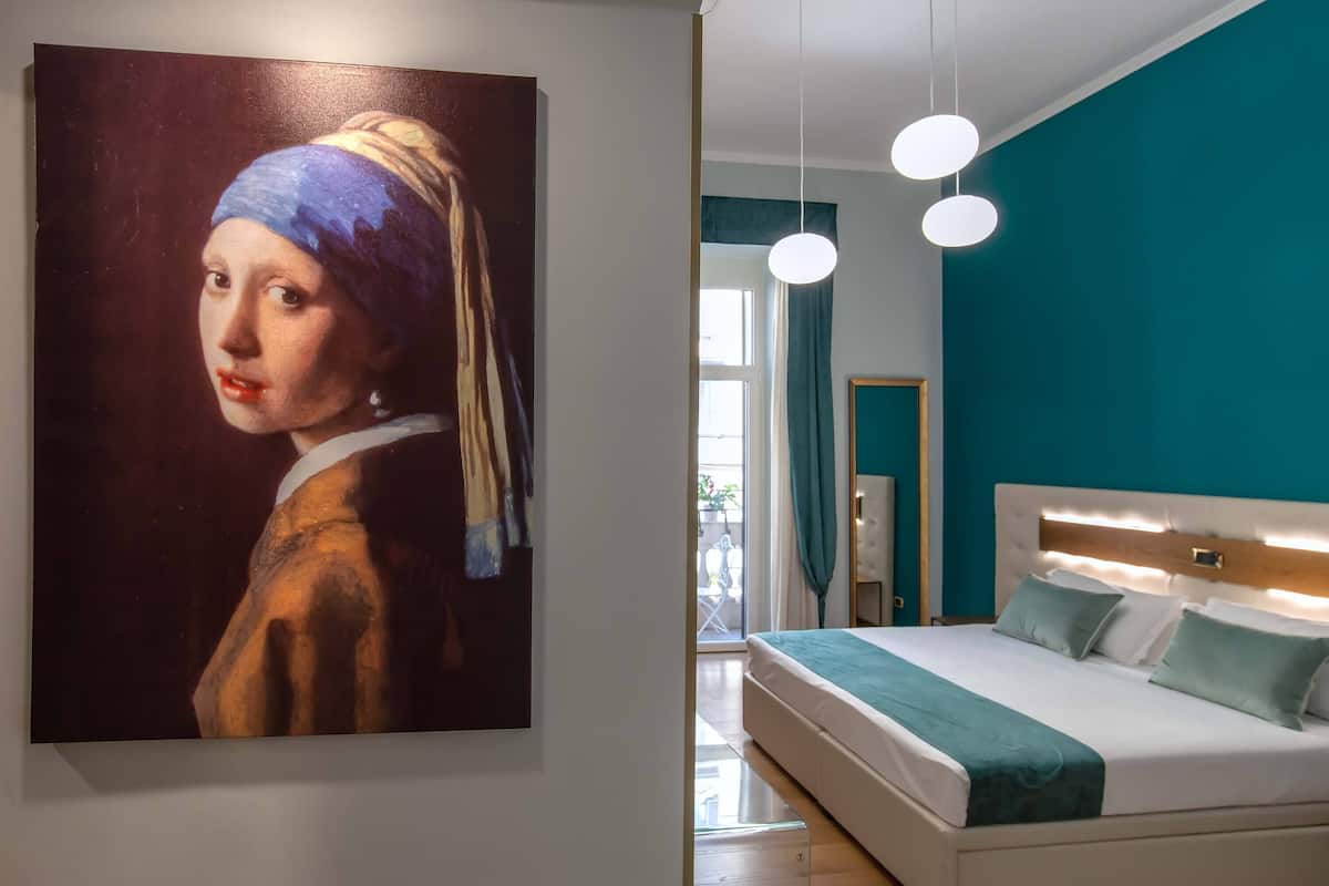 Un dormitorio con un cuadro grande, una cama con ropa de cama blanca, paredes color turquesa y lámparas colgantes.