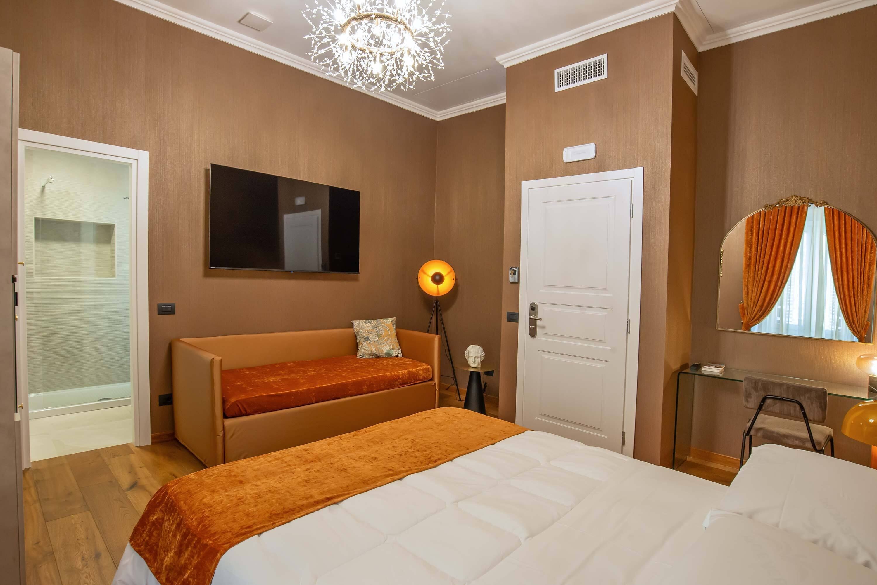 Habitación de hotel con cama, sofá, televisión, puerta, espejo y lámpara.