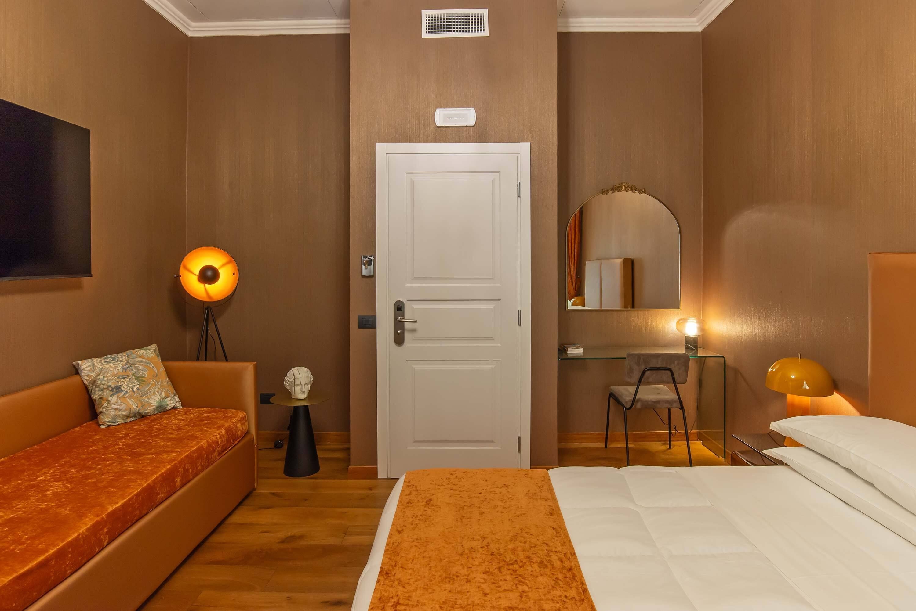 A hotel room with a bed, a sofa, a desk, a chair, a TV, and a mirror.
