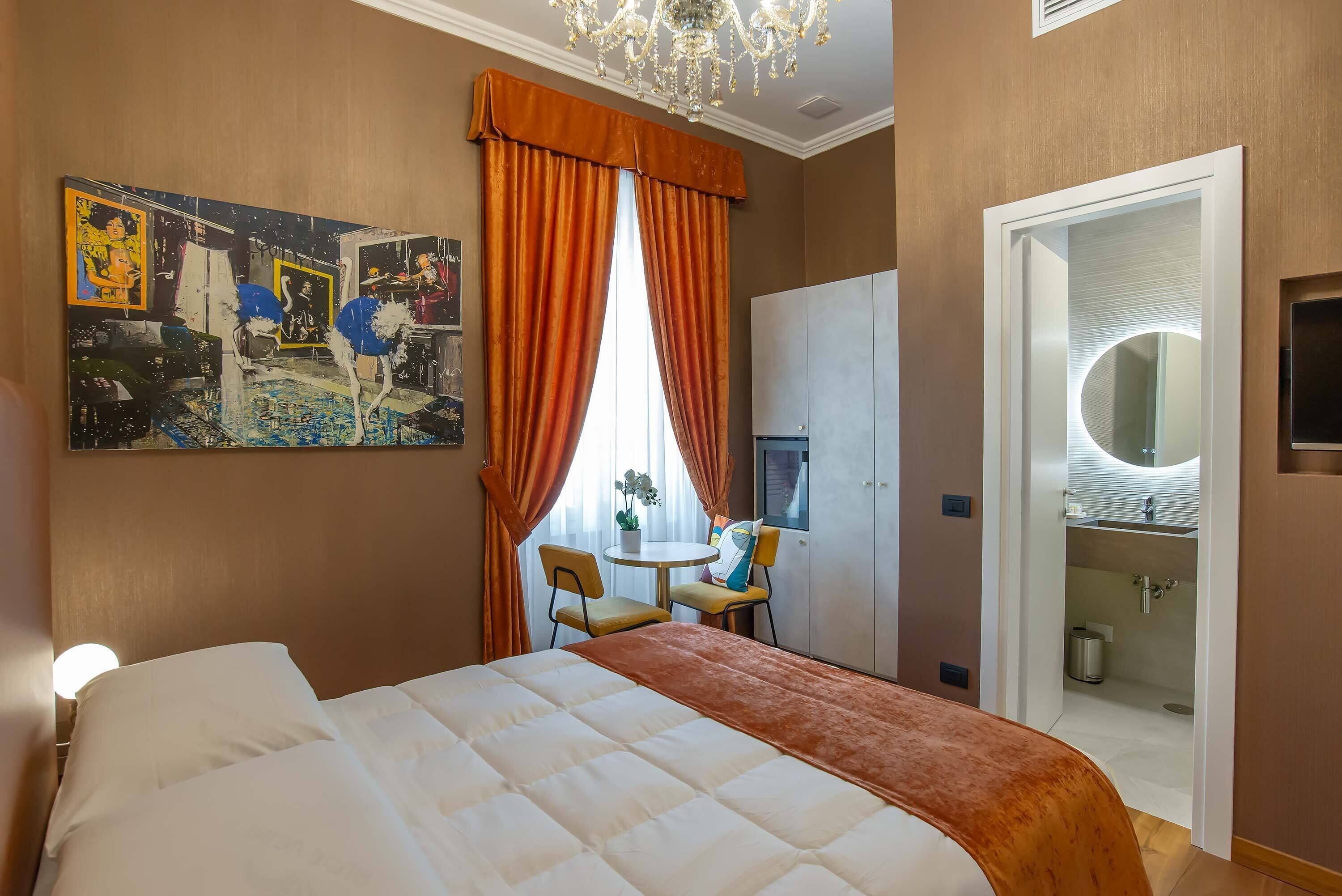 A hotel room with a bed, a painting on the wall, a desk with chairs, and a bathroom visible through an open door.