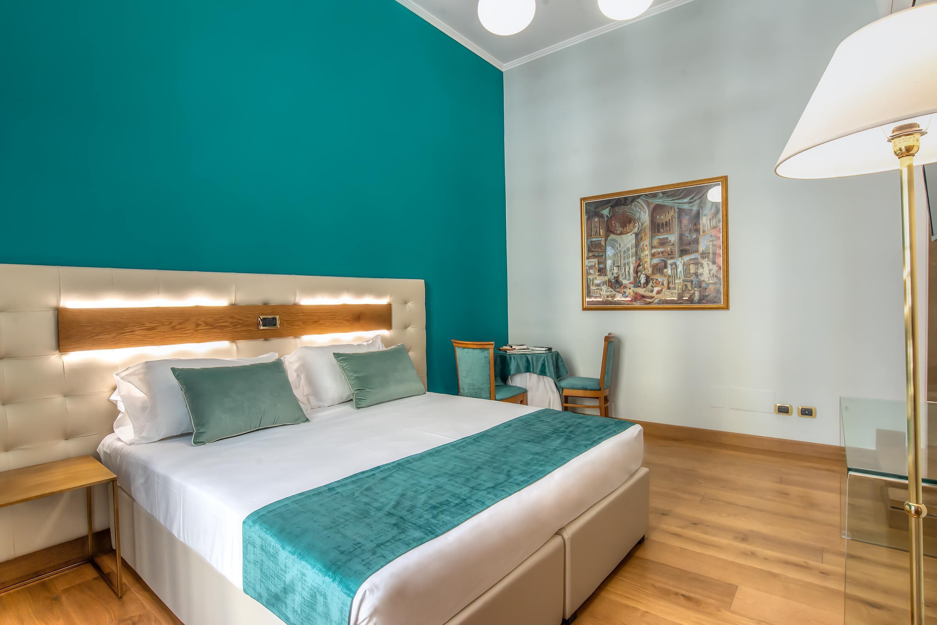 A modern bedroom with a teal accent wall, a large bed with a wooden headboard, a bedside table, a chair, a small table with a lamp, and a painting on the wall.