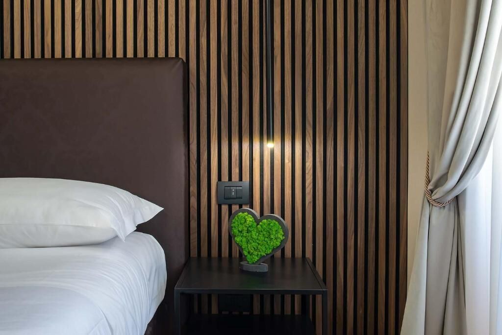 A neatly made bed with white linens, a small bedside table with a green heart-shaped decoration, and a wall with vertical wooden slats.