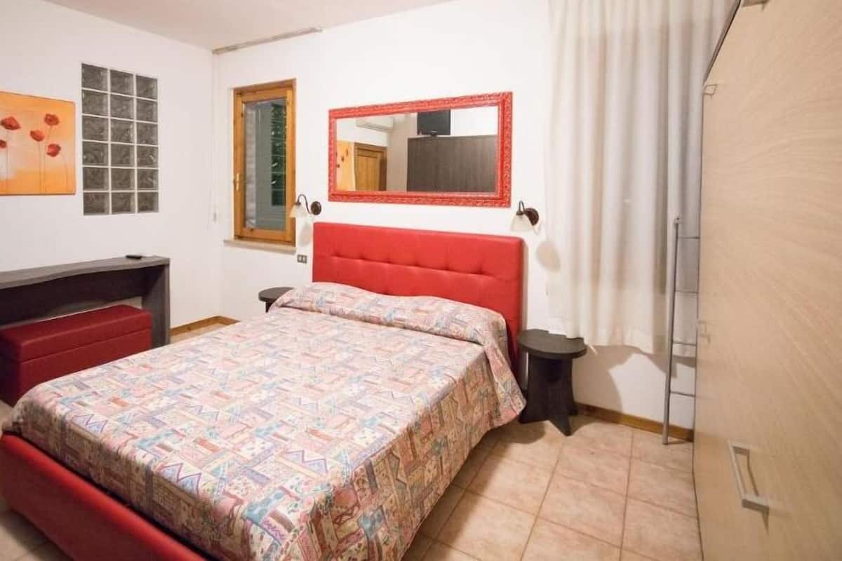 A bedroom with a red bed, a small bedside table, a mirror, a window with blinds, and a wall-mounted TV.