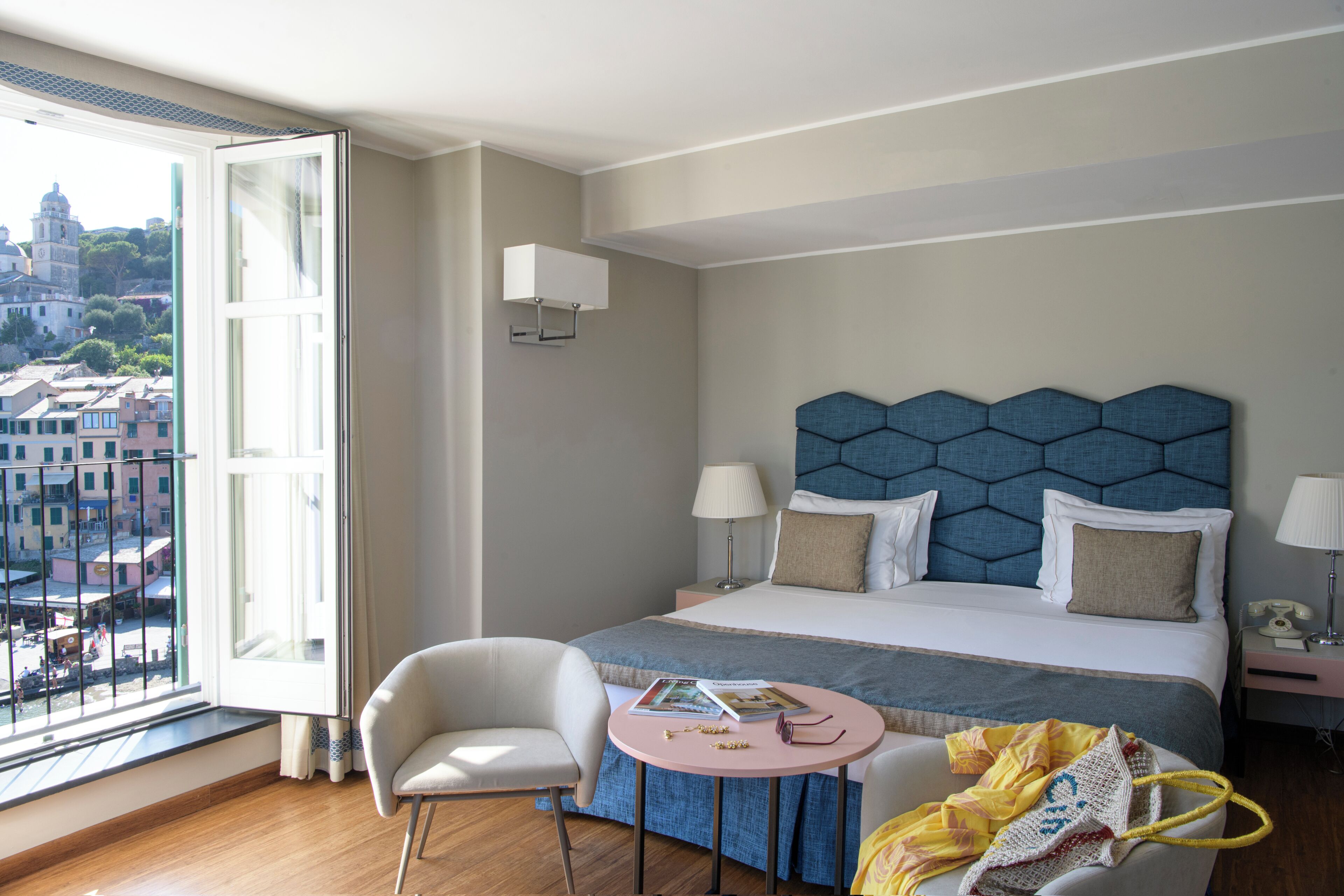 A hotel room with a large bed, a round table, a chair, and a view of the city through an open window.