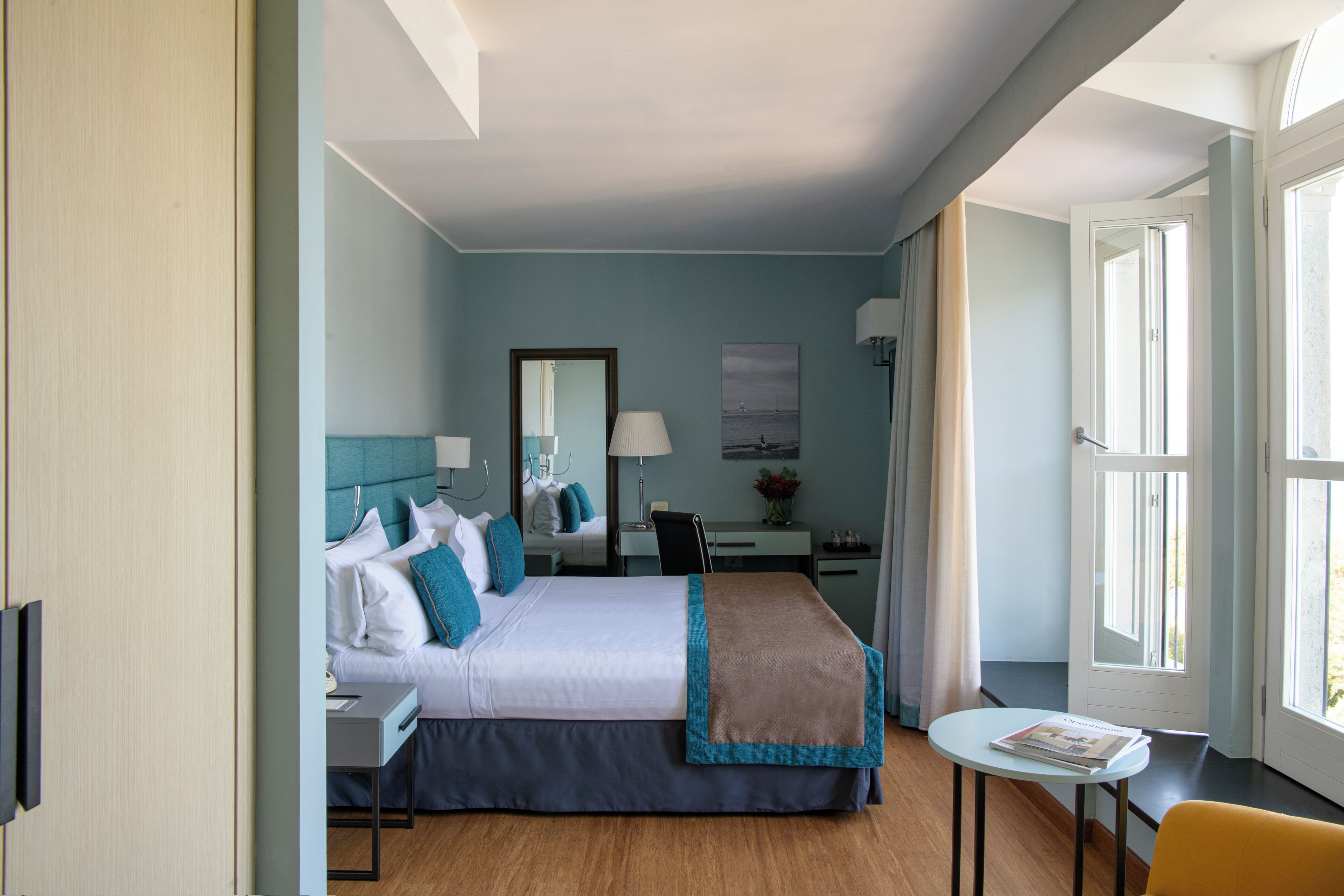 A hotel room with a bed, bedside table, chair, desk, lamp, and a window with curtains.